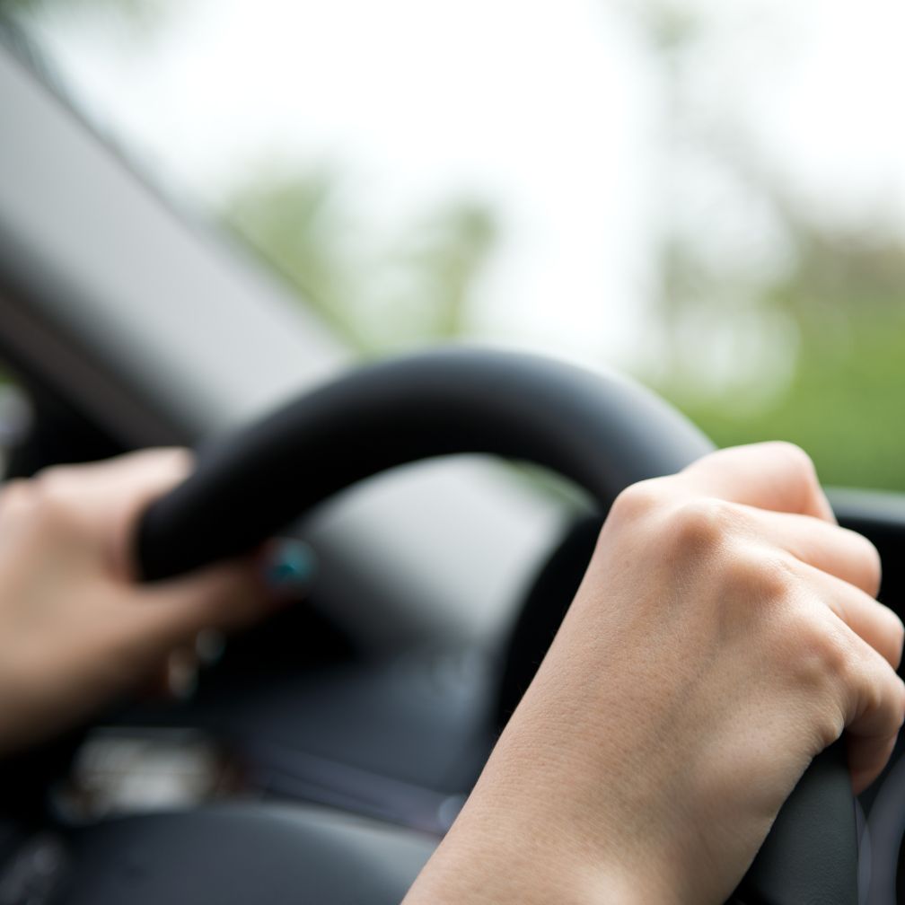 Close-up of a person’s hands gripping a black steering wheel while driving a car. The background is blurred, showing green outdoor scenery through the window.