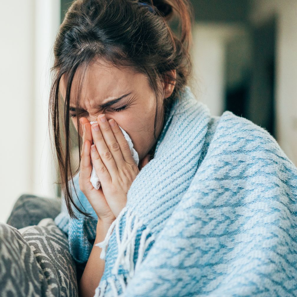 A woman wrapped in a blue blanket is sitting on a couch, holding a tissue to her nose and appearing to sneeze or cough.