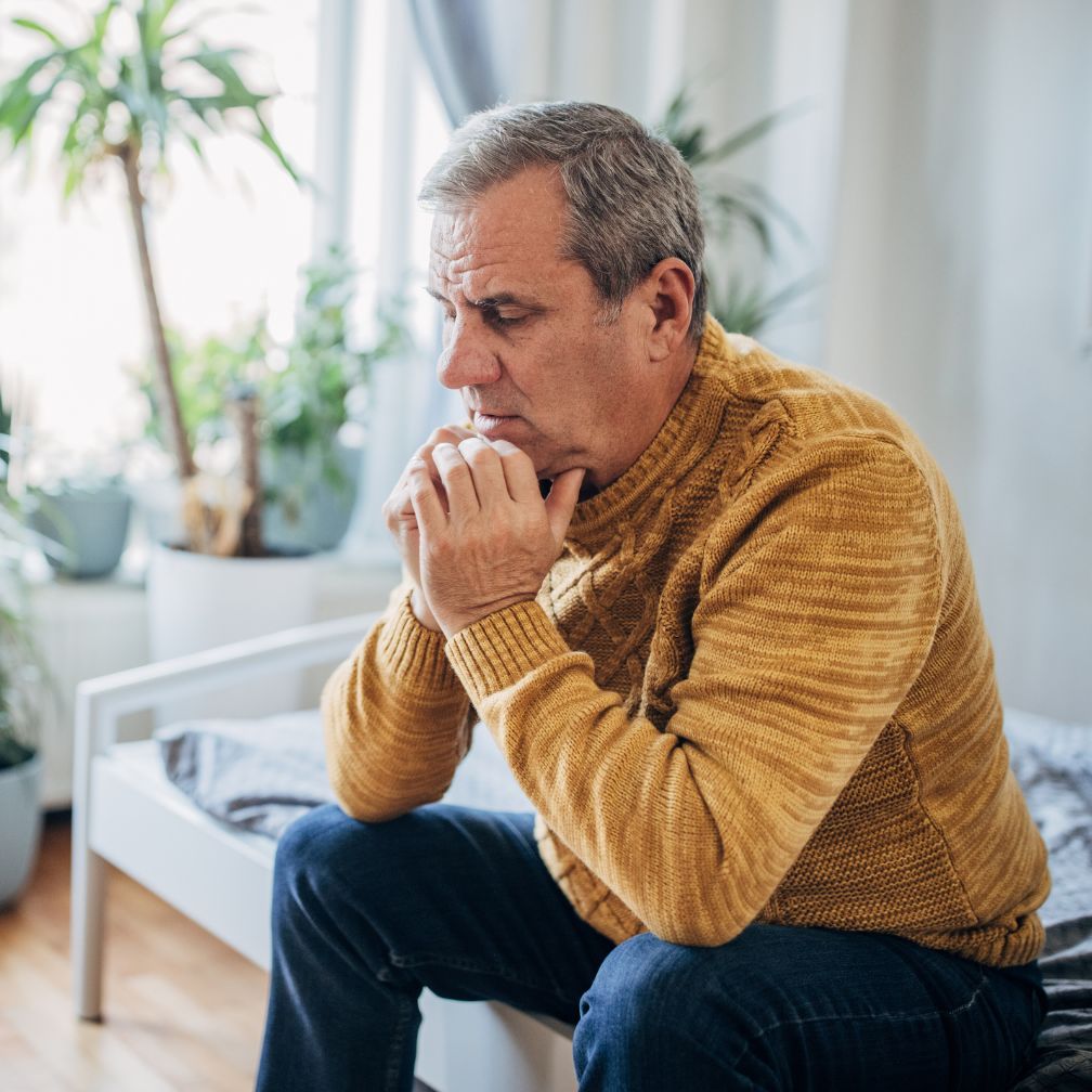 An older man in a yellow sweater sits on a bed, looking pensive and worried, with his hands clasped under his chin in a bright room filled with plants.