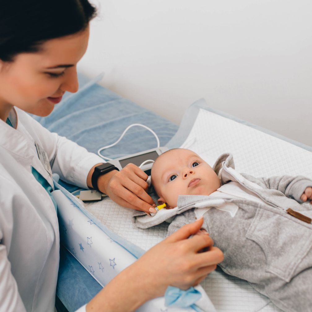 A healthcare professional tests a babys hearing using earphones while the baby lies on an exam table, looking up. The environment is clean and clinical.