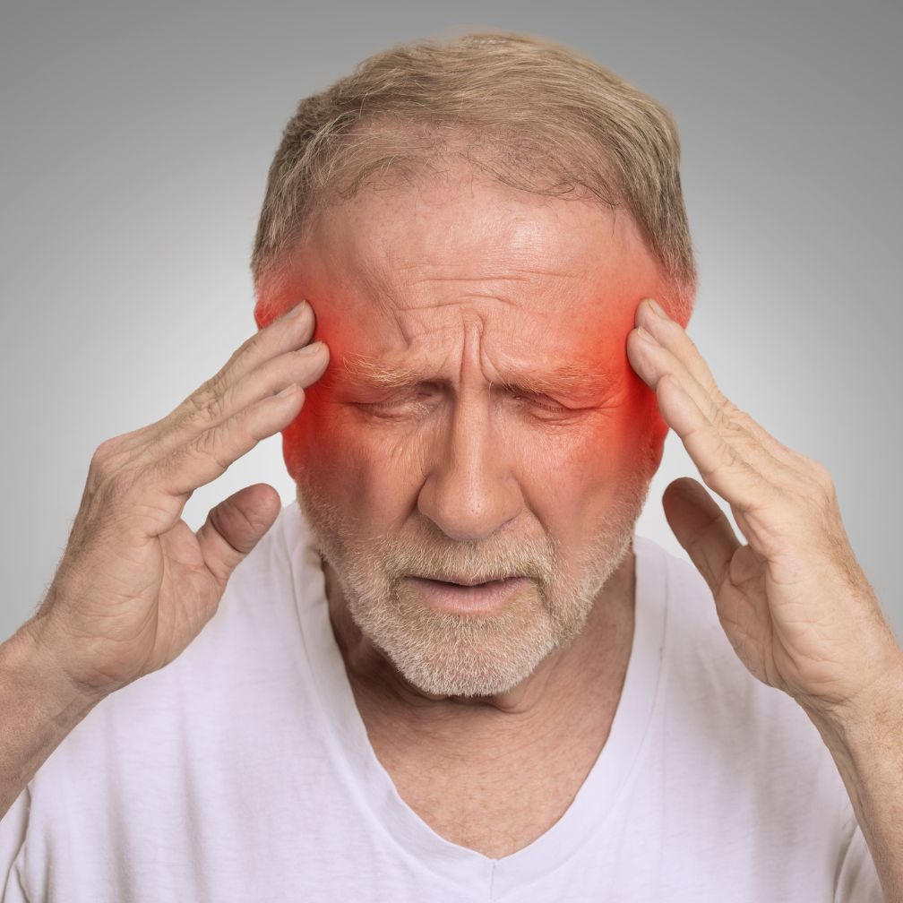 An older man with gray hair touches his temples with both hands and closes his eyes, showing pain, with red coloring on his temples to highlight a headache or migraine.