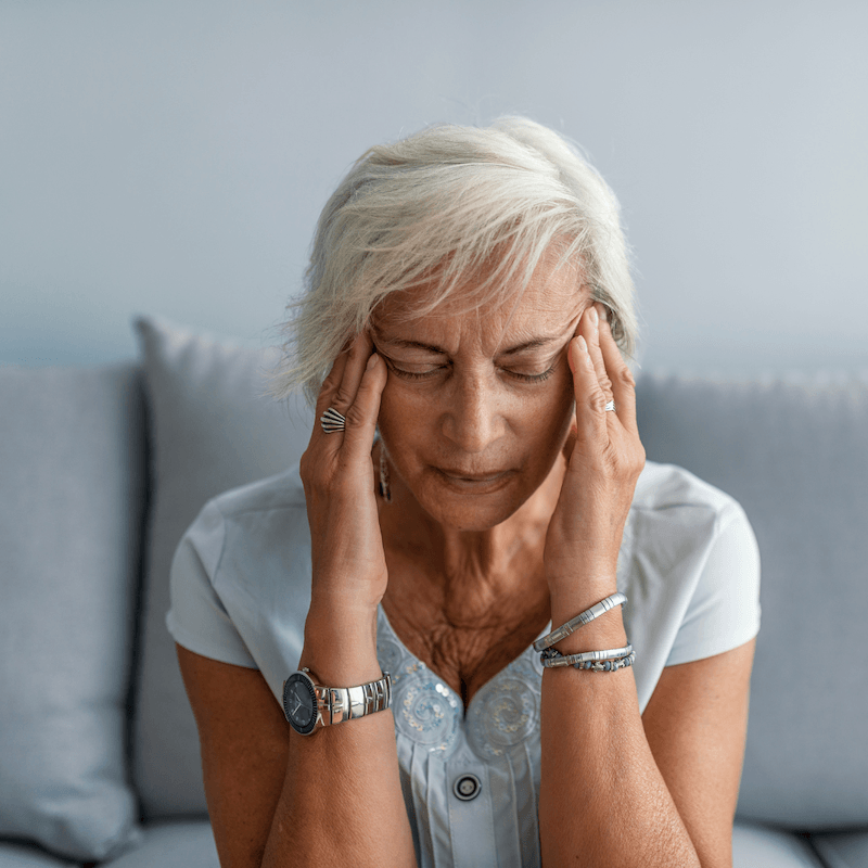 An older woman with short gray hair sits on a couch with her eyes closed, pressing her temples with both hands as if experiencing a headache or stress.