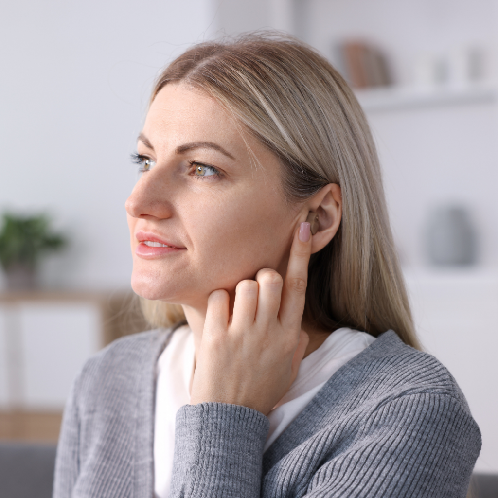 A woman with blonde hair touches her ear while wearing a hearing aid, looking slightly to the side. She is indoors, dressed in a gray sweater, with shelves and a plant blurred in the background.