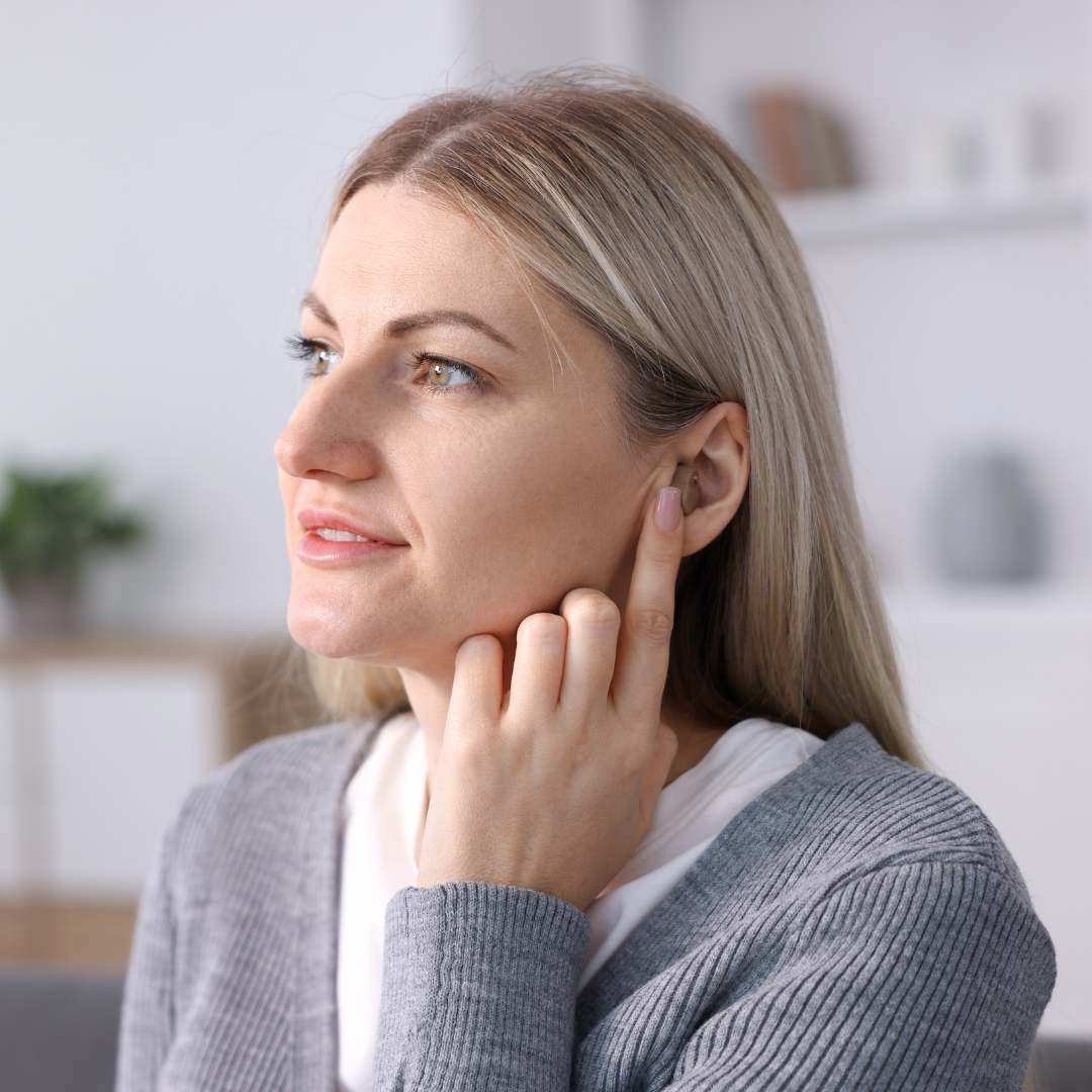 A woman with blonde hair touches her ear while wearing a hearing aid, looking slightly to the side. She is indoors, dressed in a gray sweater, with shelves and a plant blurred in the background.