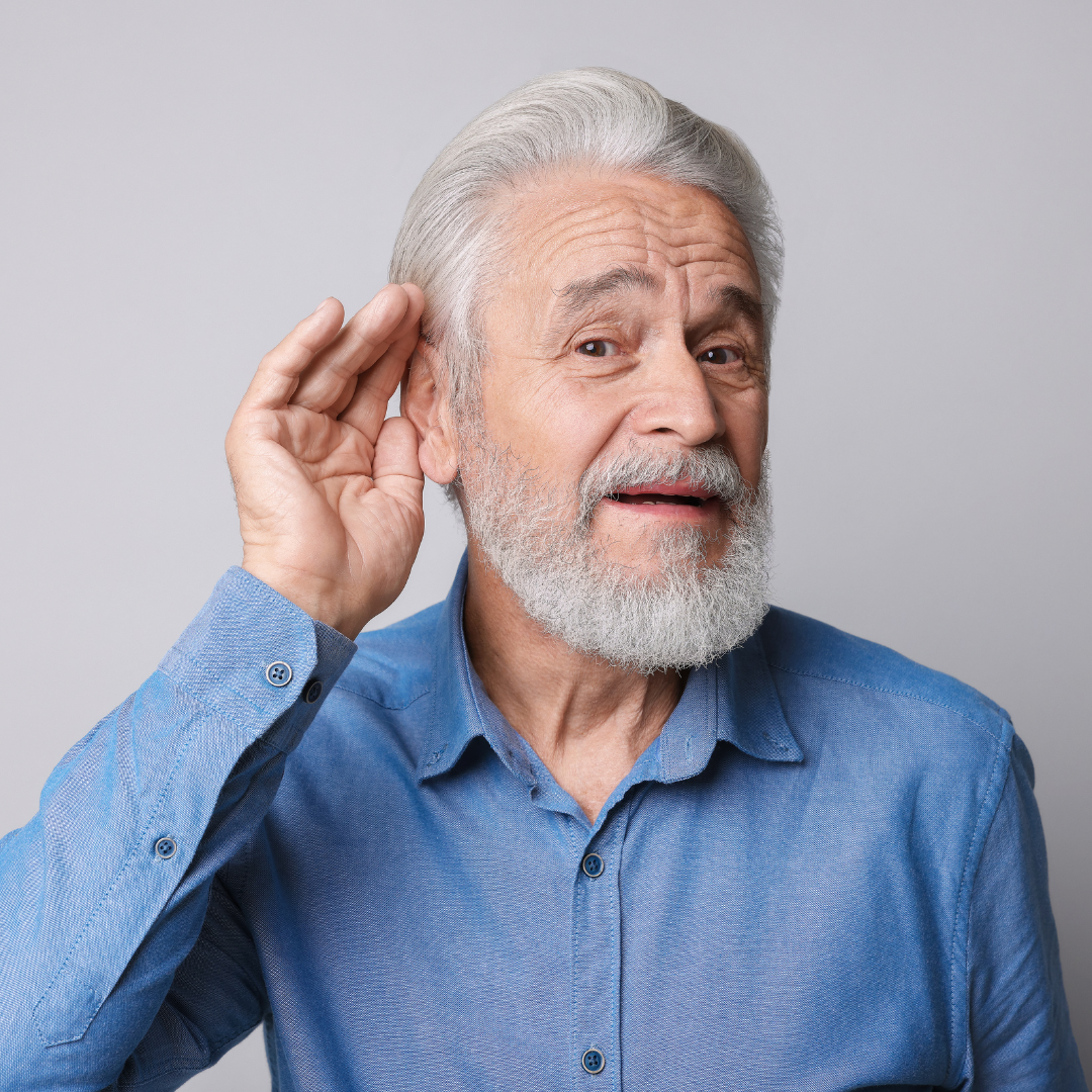 Older man with gray hair and beard wearing a blue shirt, holding his hand to his ear and leaning forward as if trying to hear better, standing against a plain light gray background.