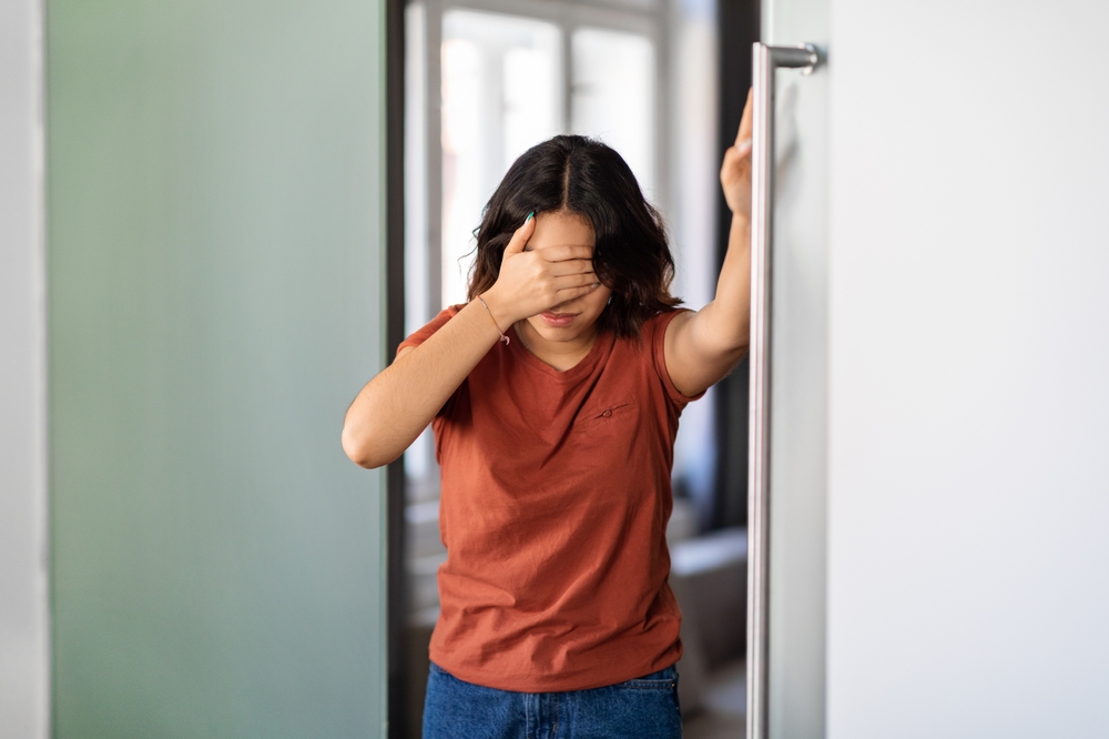 A person in a rust-colored t-shirt leans against a door frame with their hand over their eyes.