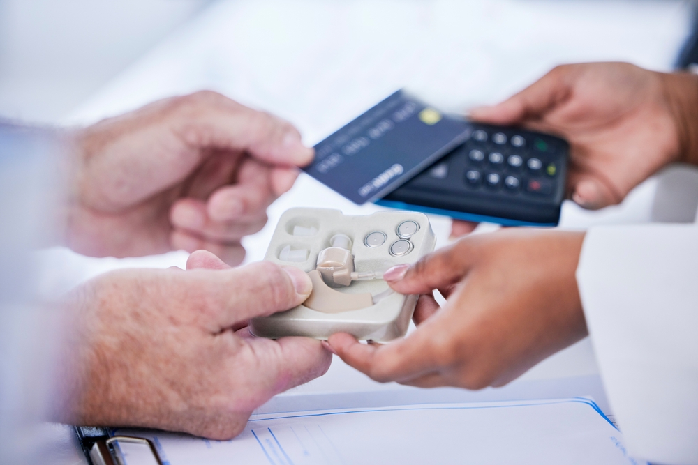 A person pays with a credit card at a terminal while receiving a hearing aid from a healthcare professional.